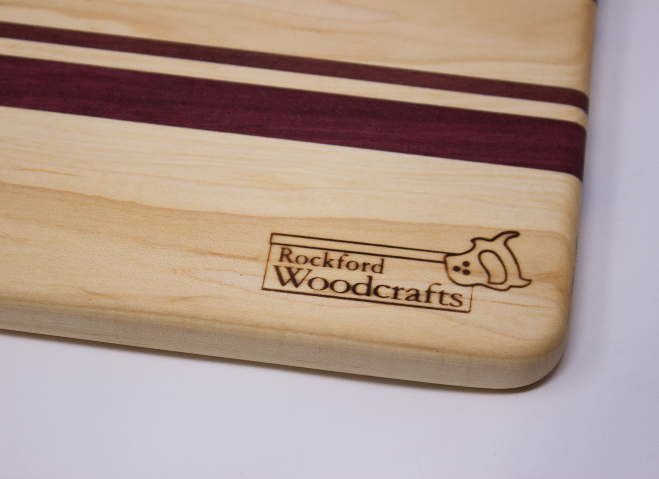 https://www.rockfordwoodcrafts.com/wp-content/uploads/Mapleand-Purpleheart-Cutting-Board-with-Handle-Back-Corner-Logo-Close-Up.jpg